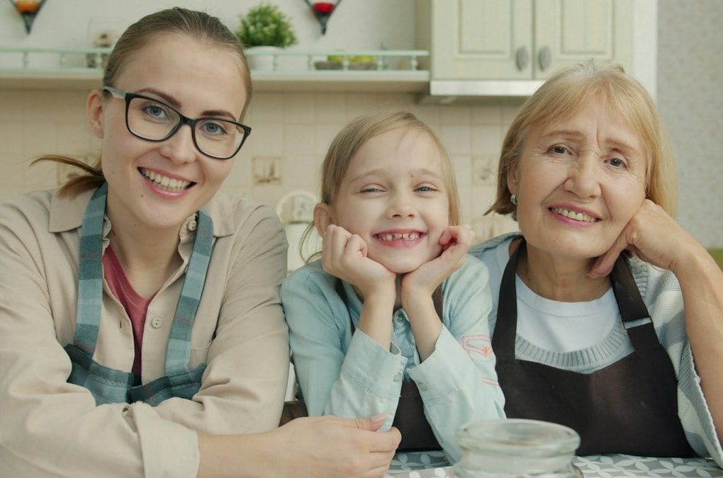 Portrait of joyful family grandmother, mother and daughter smiling in modern kitchen sitting together and looking at camera with happy faces.