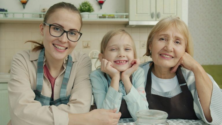 Portrait of joyful family grandmother, mother and daughter smiling in modern kitchen sitting together and looking at camera with happy faces.