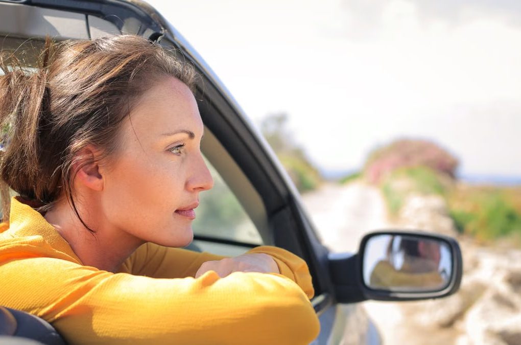 Profile view of a woman leaning out a convertible car enjoying a sunny day on a scenic road.